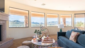 A living room with a view of a desert landscape through large windows, a fireplace, a blue sofa, and a round coffee table with various items. - Luxe Scottsdale Desert Oasis Infinity Pool, Spa, Panoramic Views + Firepit (Cave Creek)