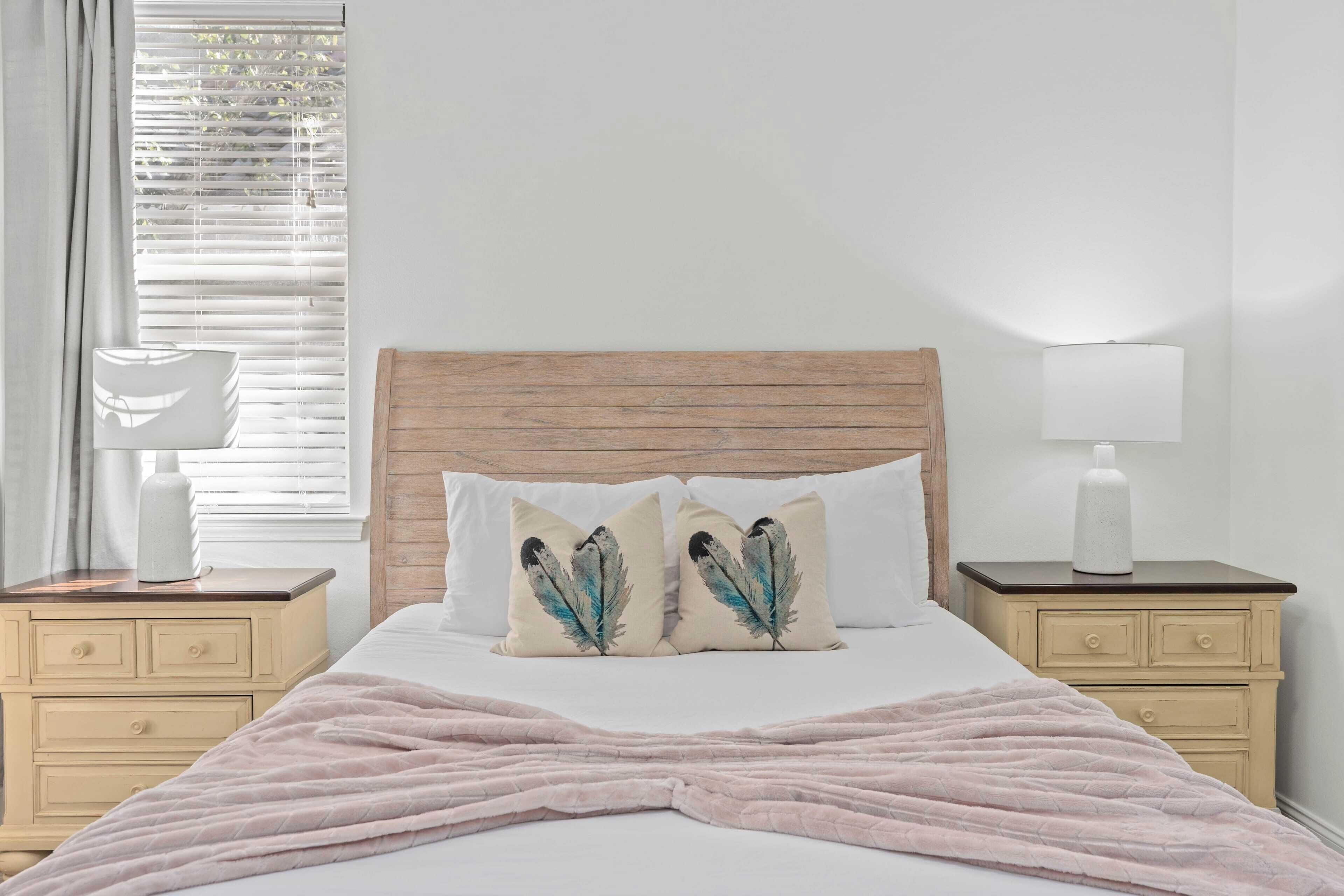 Une chambre à coucher avec une tête de lit en bois, deux tables de chevet avec des lampes, un lit recouvert d’une couverture rose et une fenêtre avec des stores.