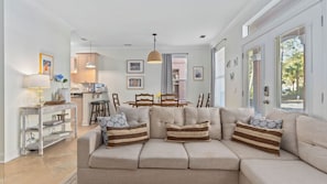 A living room with a sofa, a dining table, chairs, a kitchen area, and a hanging light fixture.