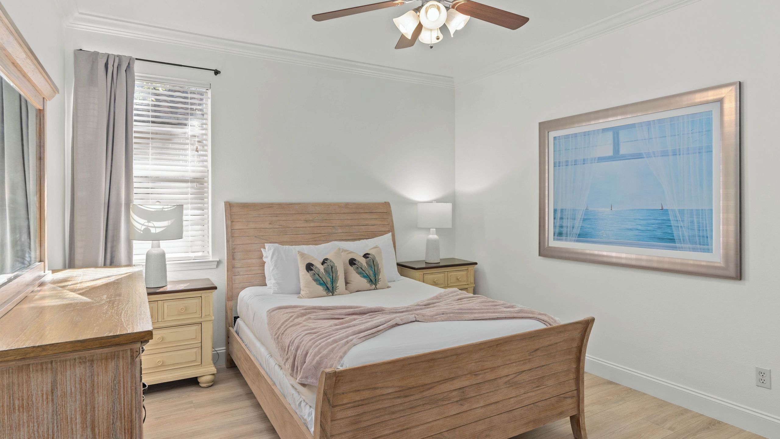A bedroom with a wooden bed, bedside tables, a ceiling fan, a window with curtains, and a framed picture on the wall.