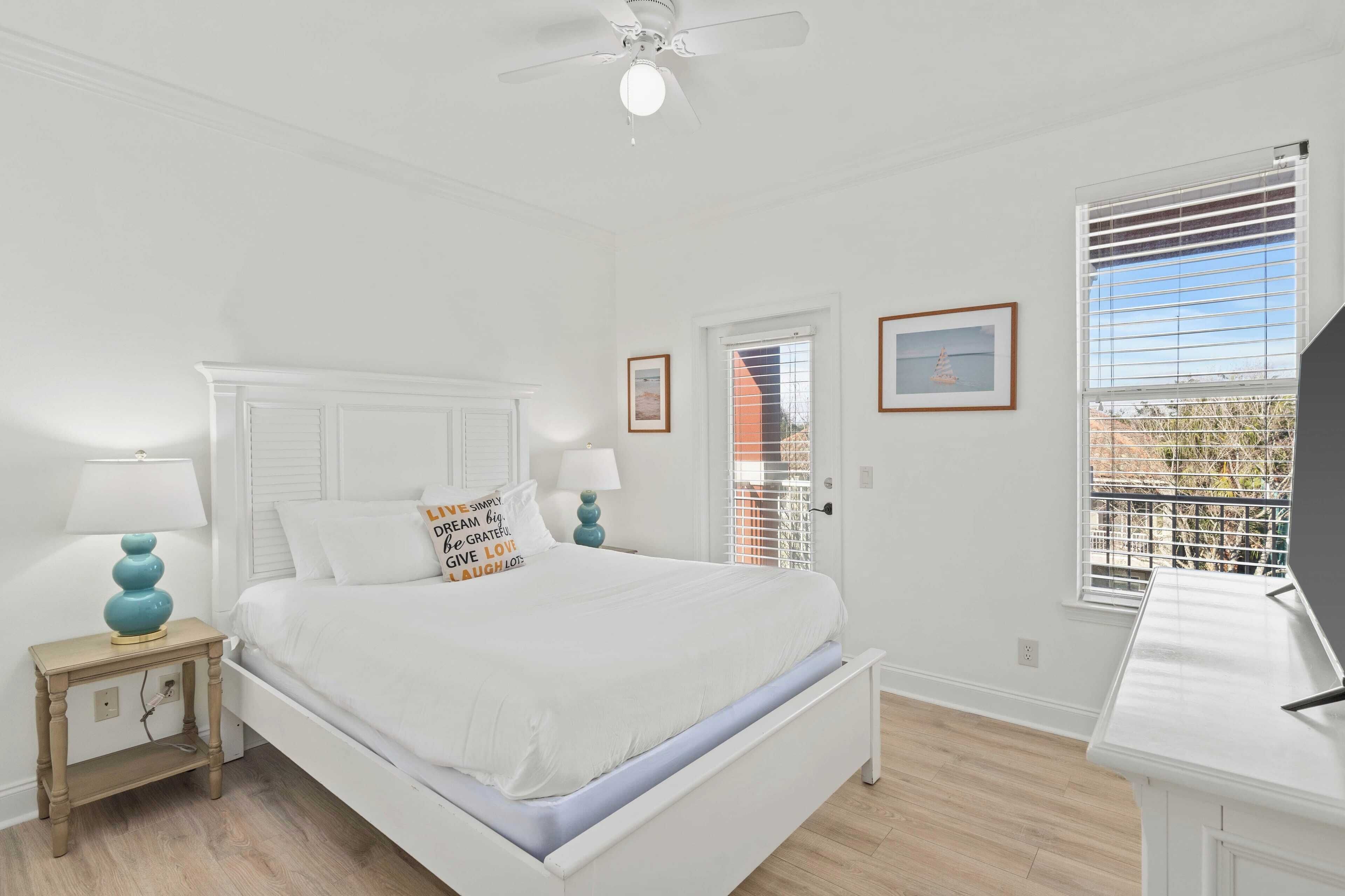 Un dormitorio con una cama blanca, dos mesitas de noche con lámparas, un ventilador de techo y una ventana con persianas.