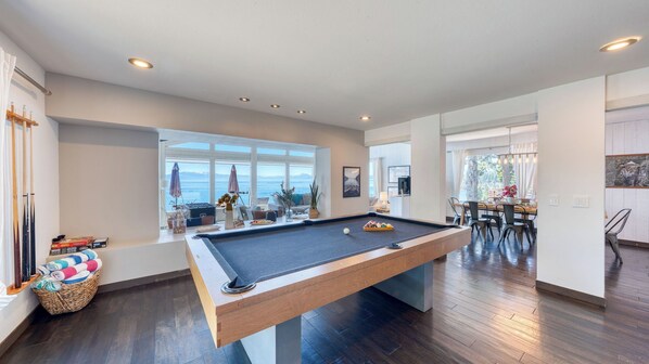 A pool table in a modern living room with a view of the sea, a dining area, and a kitchen.