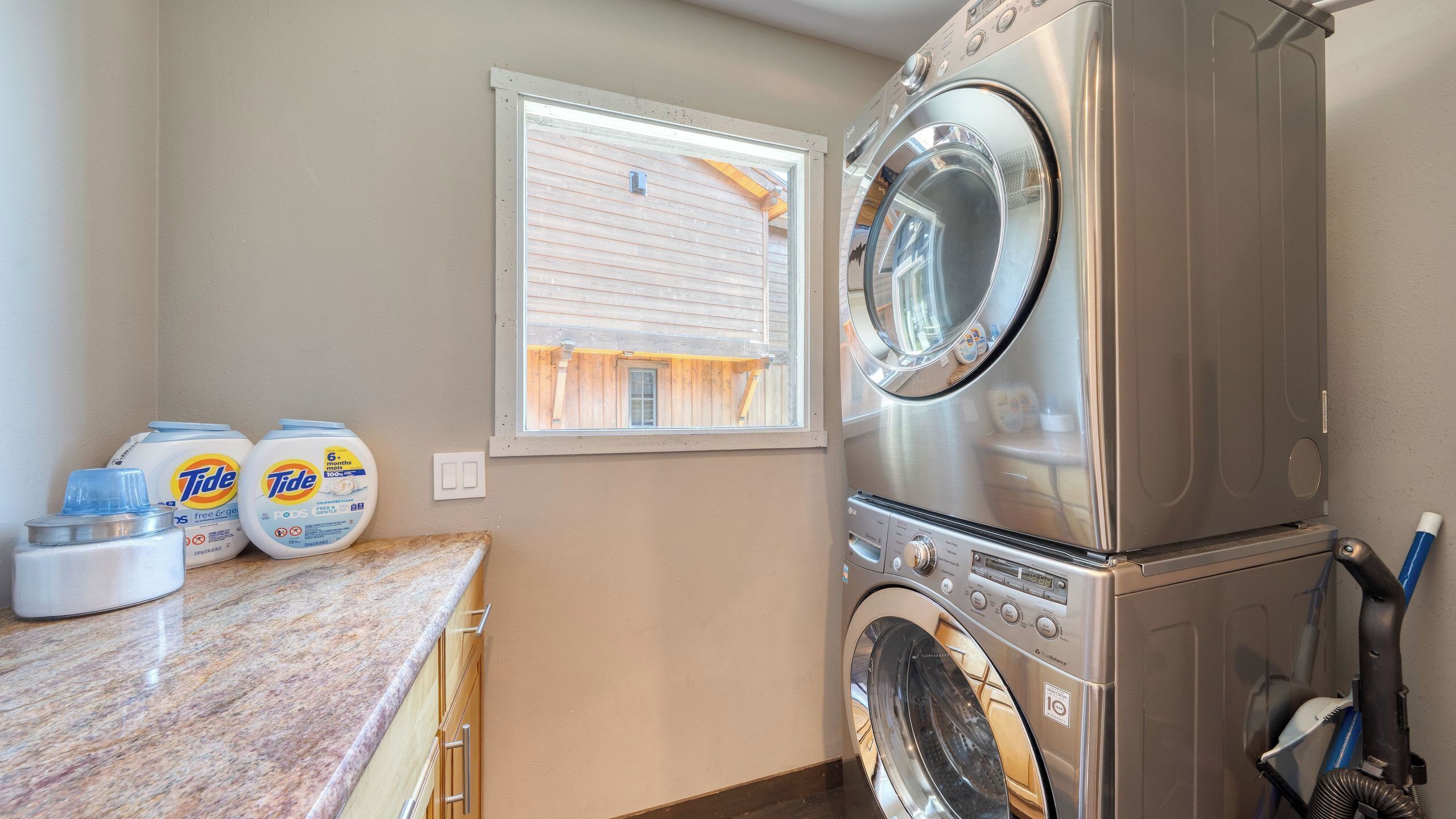 A laundry room with a stacked washer and dryer, Tide detergent, and a window with a view of a wooden building.