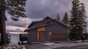 A modern two-story house with a garage, surrounded by trees and a body of water.