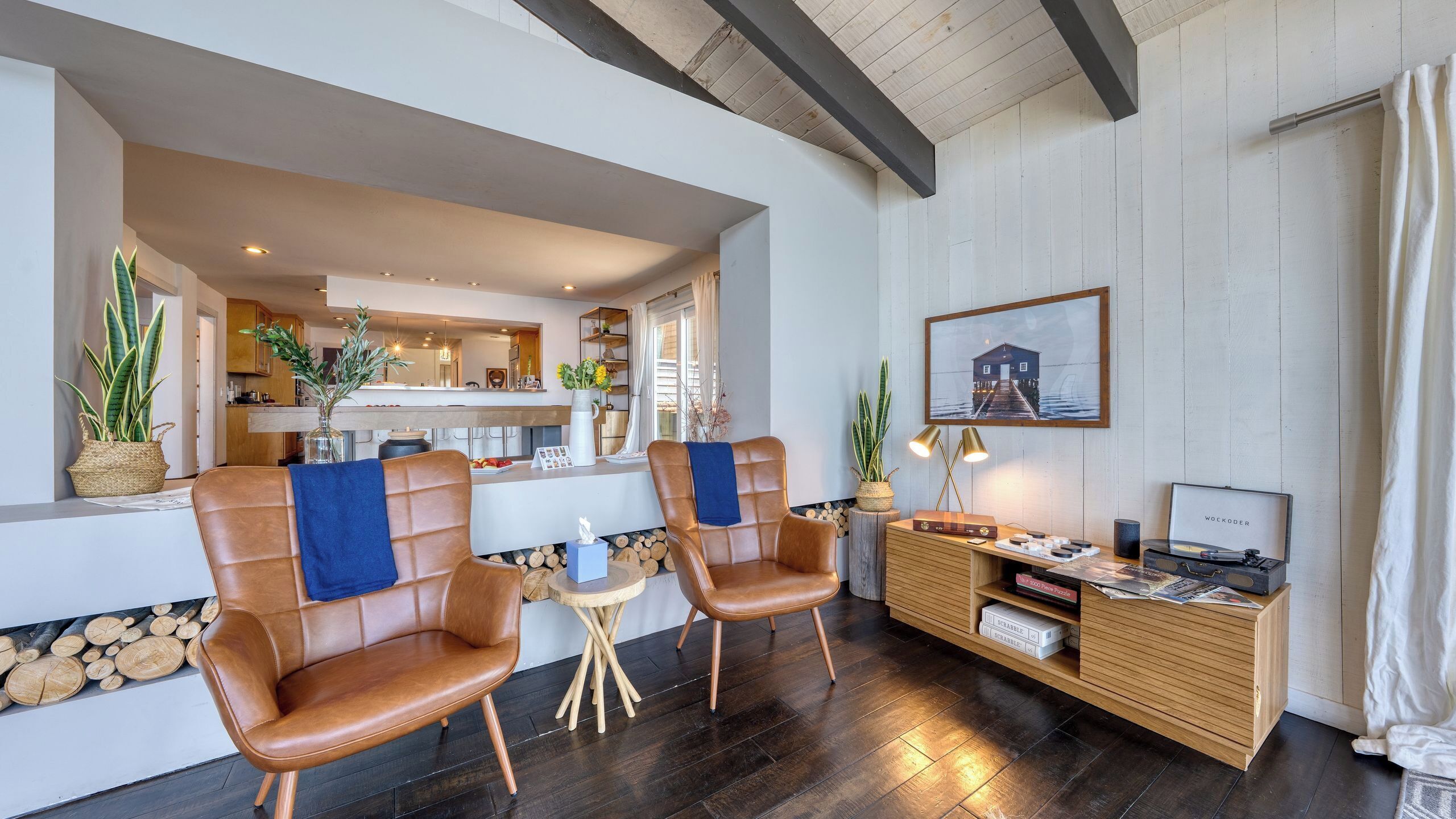 A modern living area with leather chairs, a wooden console table with board games, and a kitchen in the background.