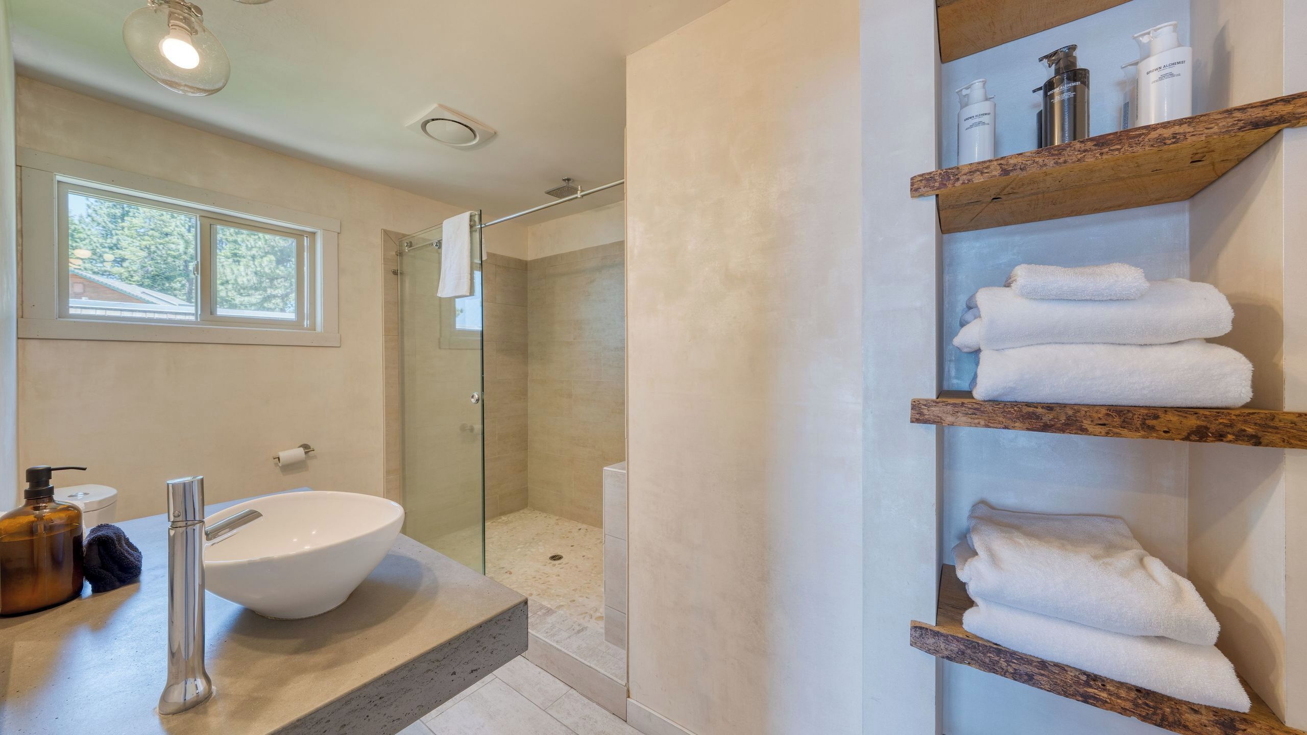 A bathroom with a large white sink, a glass shower enclosure, and wooden shelves with towels.