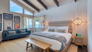 A bedroom with a large bed, a blue sofa, a wooden bench, and a view of the sea.