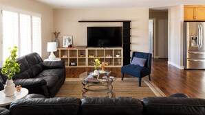 A modern living room with a black leather sofa, a glass coffee table, and a blue armchair. A flat-screen TV is mounted on the wall, and there is a kitchen area with a stainless steel refrigerator. - West Ridge by Avantstay Pool, Fire Pit, Al Fresco Dining 7 Mins to Downtown (Scottsdale)