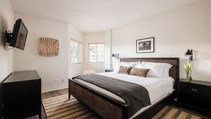 Une chambre à coucher comprenant un lit, des tables de chevet, un bureau, un téléviseur fixé au mur et une fenêtre avec des stores.