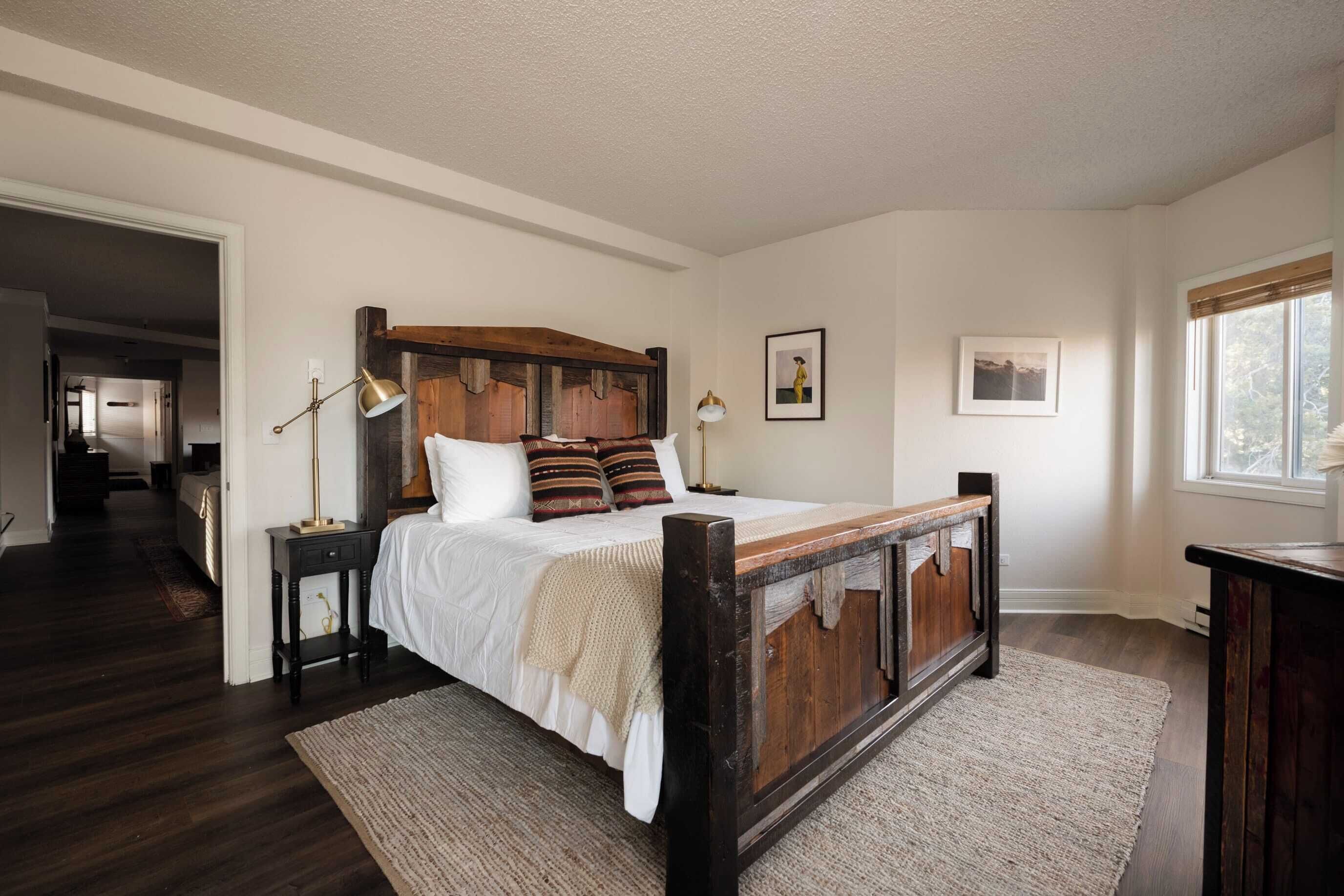 Ein Schlafzimmer mit einem großen Bett, einem Holz-Kopfteil, zwei Nachttischen, einem Fenster und einer Tür zu einem anderen Raum.