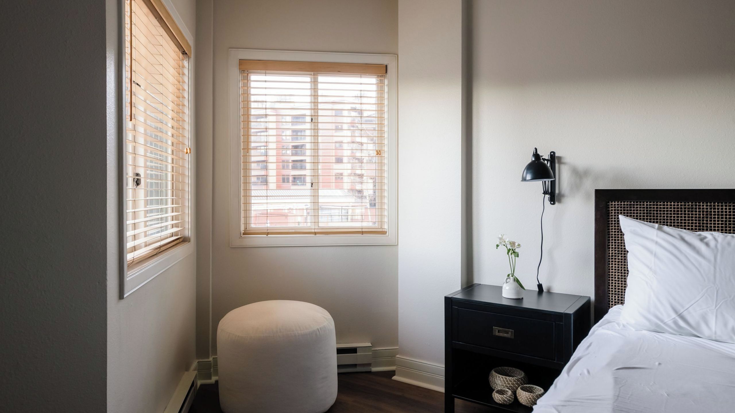 A bedroom with a bed, a nightstand with a lamp, a window with blinds, and a small ottoman.