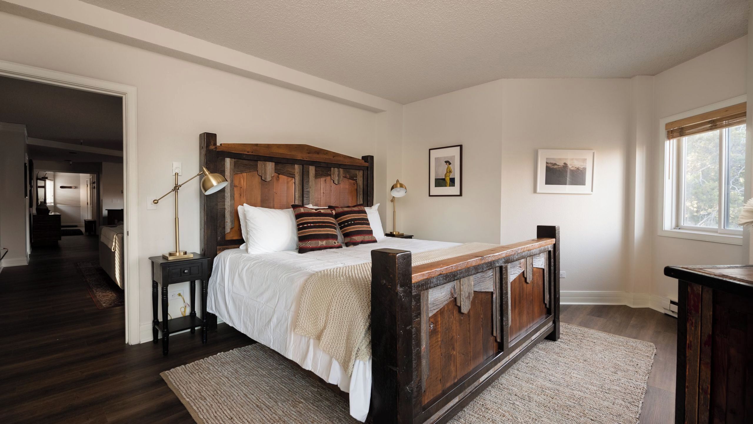 A bedroom with a wooden bed frame, white bedding, a nightstand with a lamp, and a view of the outdoors through a window.
