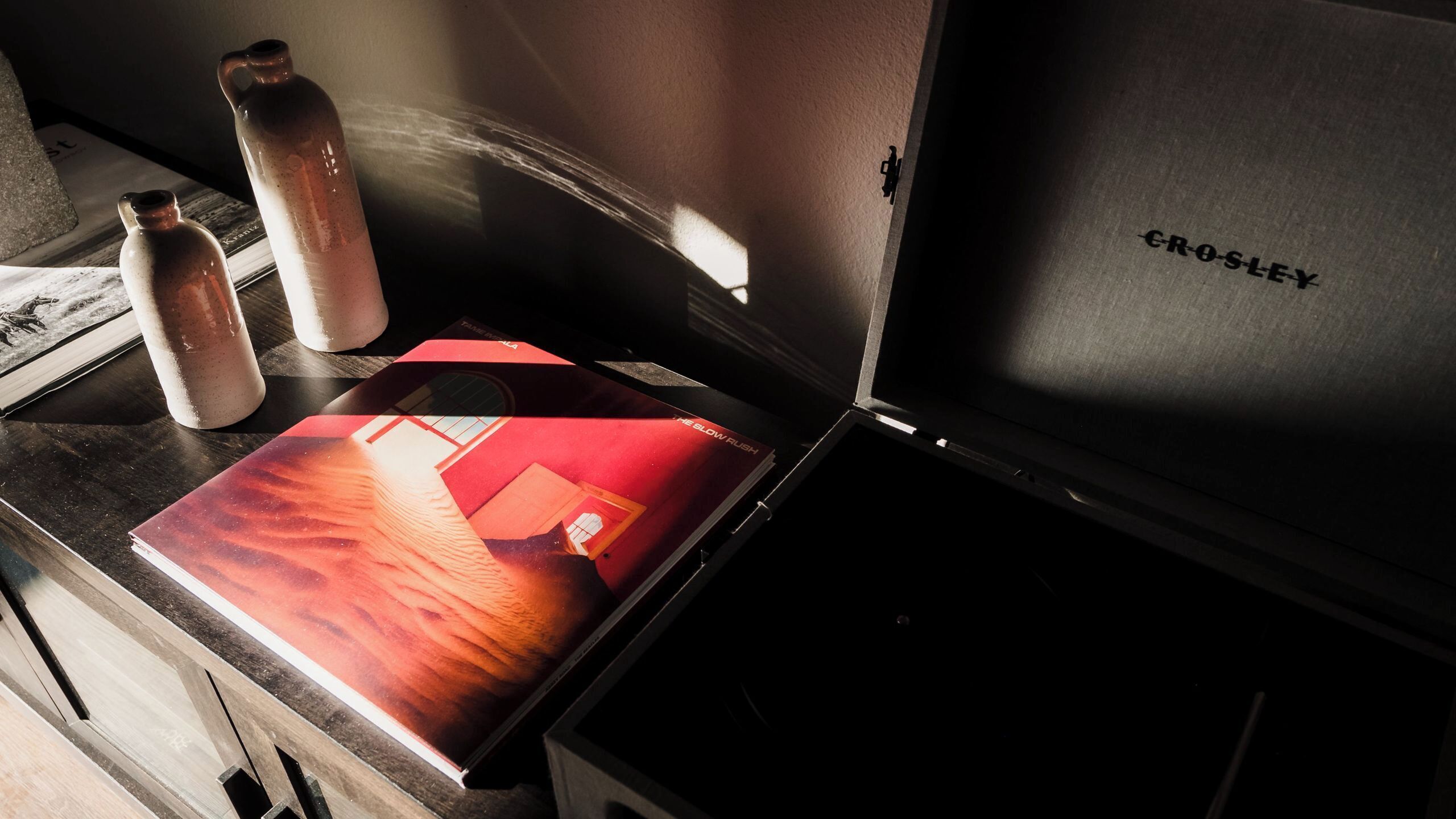 A room with a wooden desk, a red book, two ceramic water bottles, and a closed black suitcase with 'CROSLEY' text.