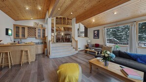 A wooden interior with a kitchen, dining area, staircase, and a sofa with a view of snow-covered trees.