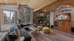 A cozy living room with a stone fireplace, wooden ceiling, and large windows overlooking a snowy landscape.