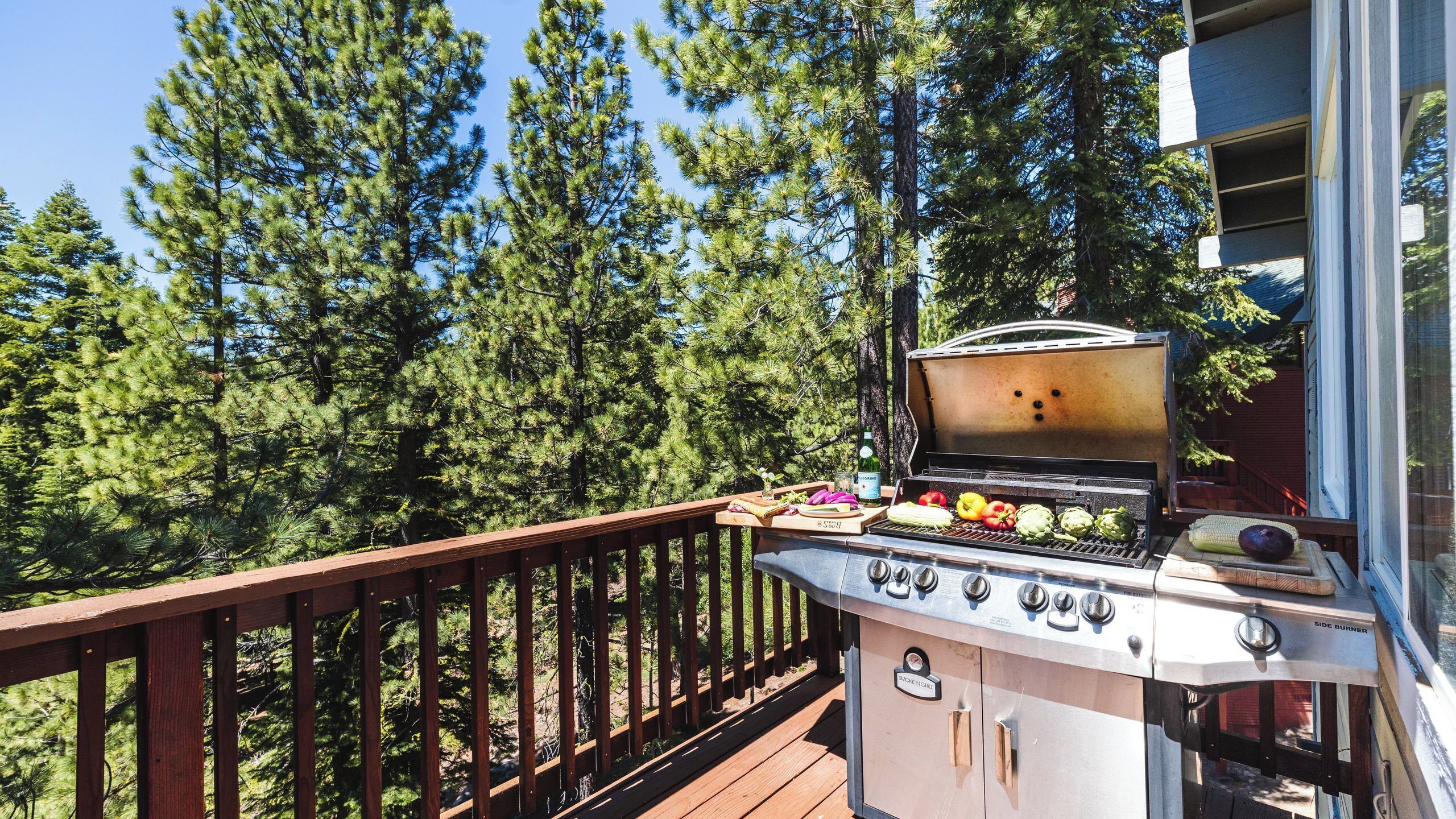 A barbecue grill on a wooden deck with a view of tall pine trees.