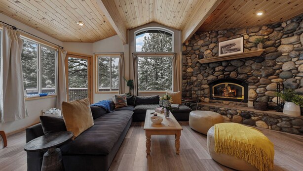 A cozy living room with a stone fireplace, large windows, and a wooden ceiling.