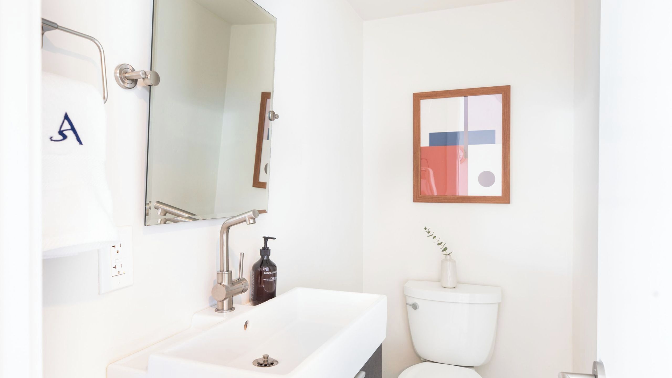 Ein Badezimmer mit einem weißen Waschbecken, einem Spiegel, einer Toilette und einem gerahmten abstrakten Kunstwerk an der Wand.