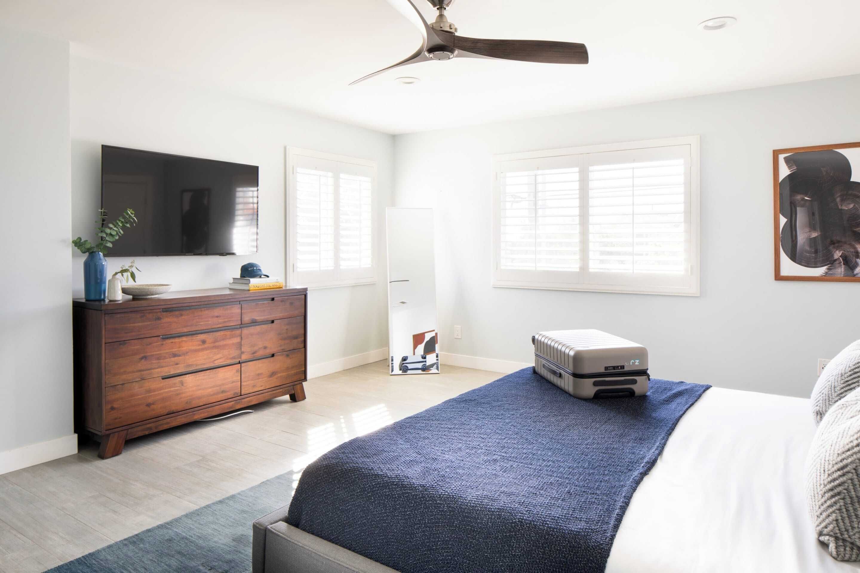 A bedroom with a bed, a dresser, a ceiling fan, a radiator, and a TV on the wall.