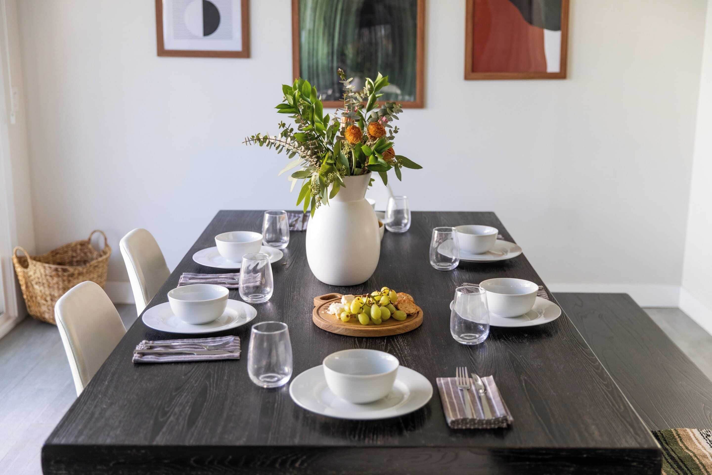 Une table à manger dressée avec de l’assiette, des bols et des verres blancs, un vase rempli de fleurs et une planche en bois sur laquelle sont disposés des raisins et du fromage.