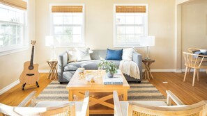 A living room with a sofa, coffee table, and chairs. There is a guitar, a lamp, and a window with a view. - Kokomo by Avantstay Bayside Getaway w/ Fire Pit, & Large Outdoor Patio (San Diego)