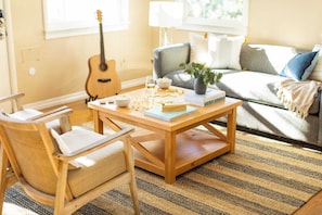 A cozy living room with a wooden coffee table, a wicker chair, a guitar, and a sofa with cushions. - Kokomo by Avantstay Bayside Getaway w/ Fire Pit, & Large Outdoor Patio (San Diego)