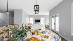 A spacious living room with a dining area, featuring a large wooden table set with food, a TV mounted on the wall, and large windows allowing natural light. - Blue Jay By Avantstay Spectacular Destin Oasis w/ Pool Walking Distance to Beach (Destin)