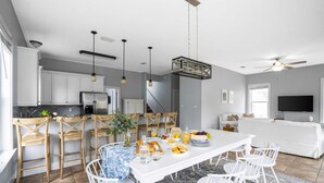 A modern kitchen with white cabinets, a dining area with a white table and chairs, and a living room with a white sofa and a flat-screen TV. - Blue Jay By Avantstay Spectacular Destin Oasis w/ Pool Walking Distance to Beach (Destin)