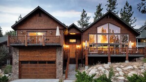 A two-story log cabin with a stone foundation, a wooden garage, and a balcony with outdoor lighting. - Bernard by Avantstay Luxurious Cabin in Big Bear w/ Hot Tub & Pool Table (Big Bear Lake)