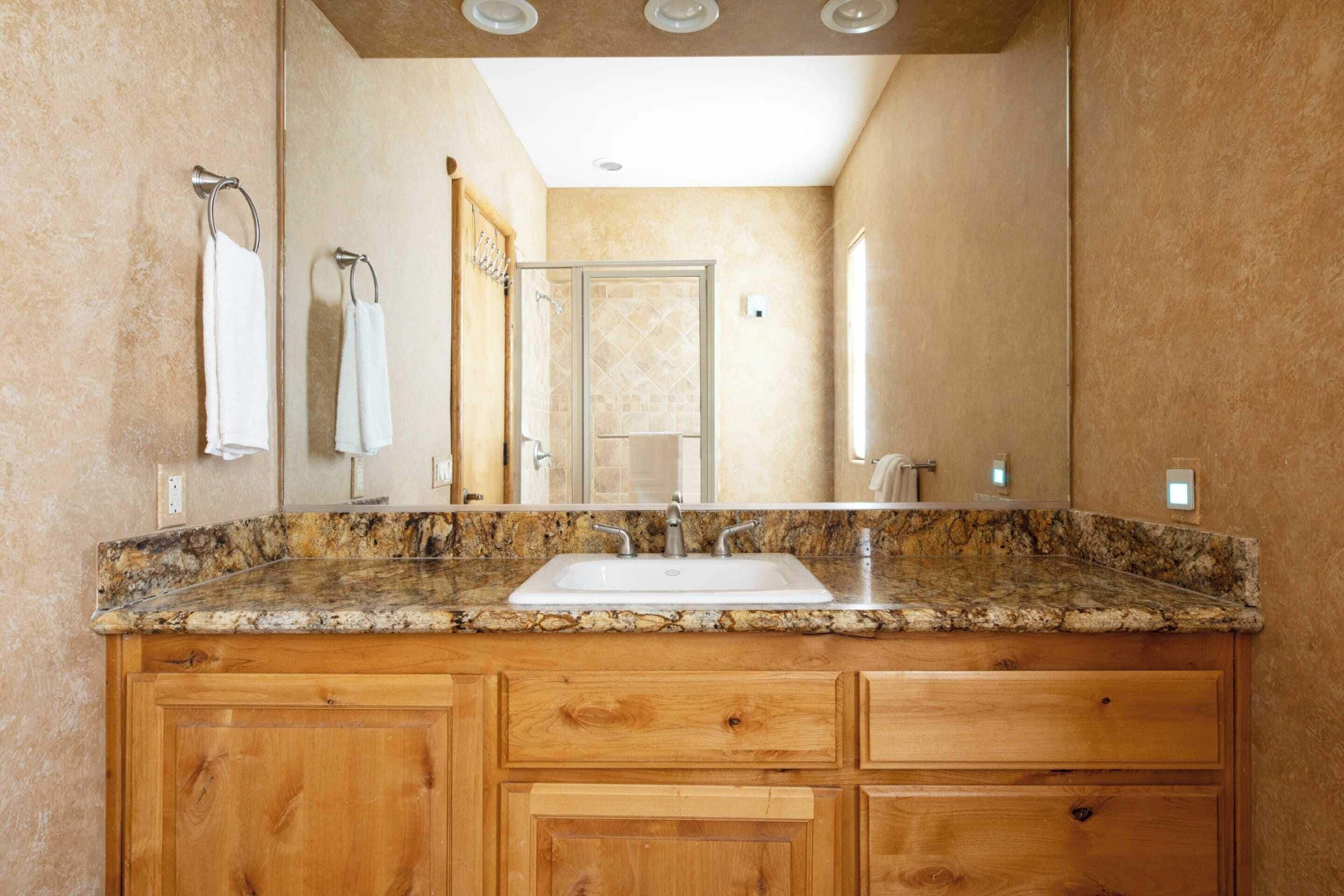 Un baño con lavamanos de encimera de granito, armarios de madera y un espejo grande.