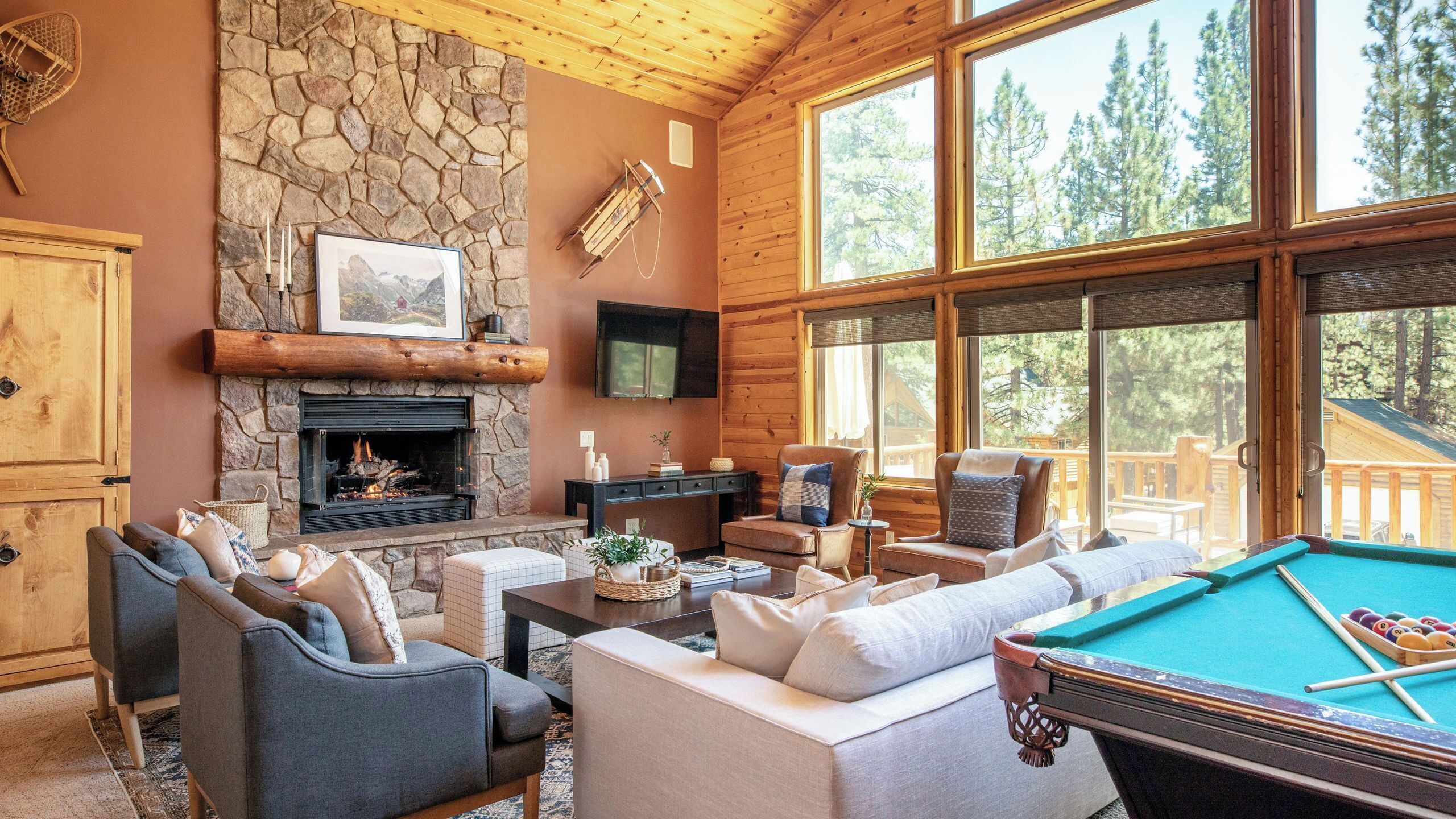 A cozy lodge interior with a stone fireplace, comfortable seating, a pool table, and large windows offering a view of the surrounding forest.
