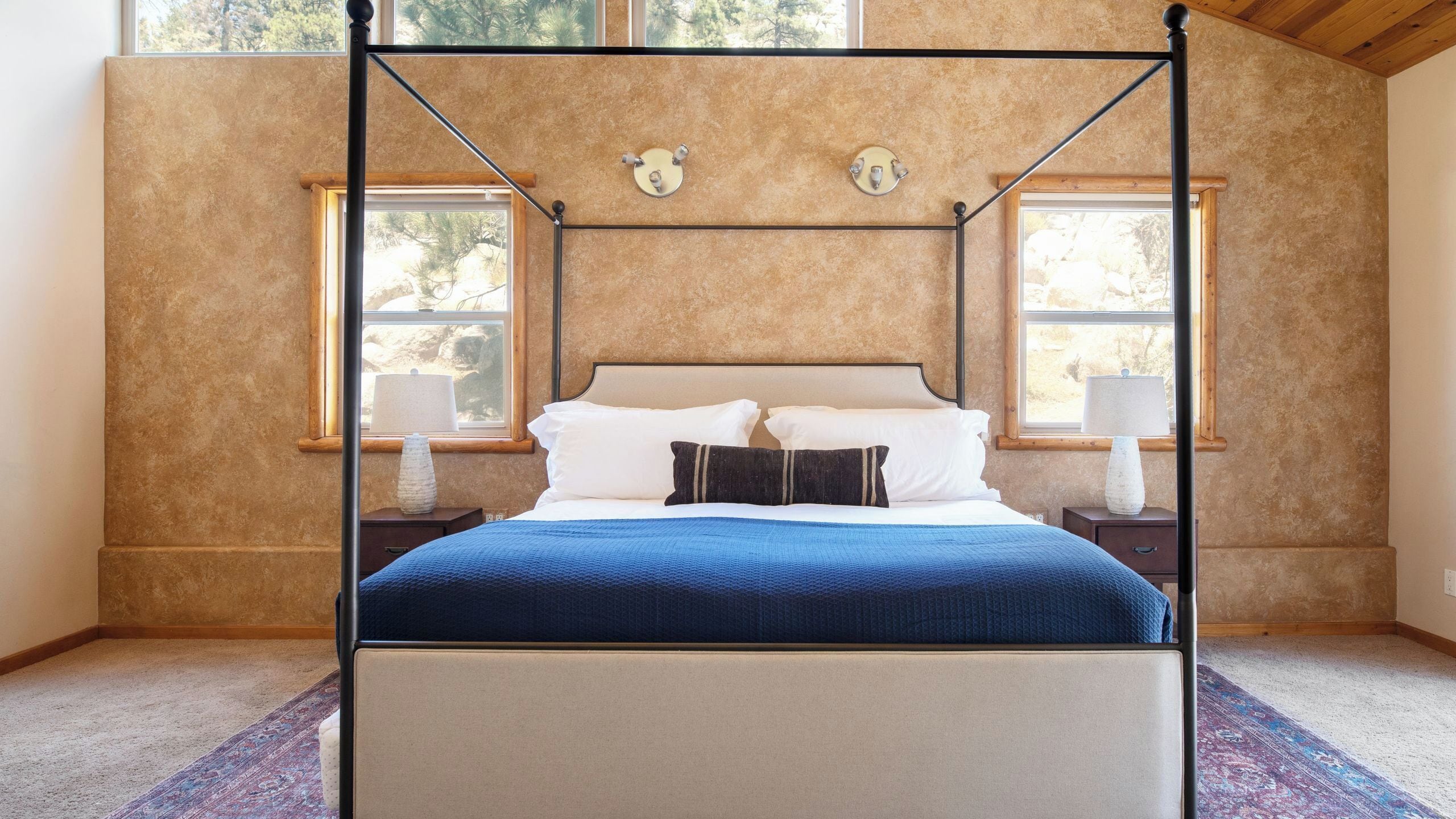 A four-poster bed with a headboard, two bedside lamps, and a window with a view of trees.