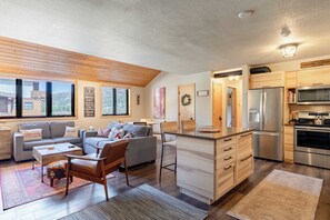 Una sala de estar moderna con un sofá, una mesa de centro y una silla de madera. Hay una zona de cocina con refrigerador, estufa y microondas. La habitación tiene ventanas amplias y vistas al exterior.