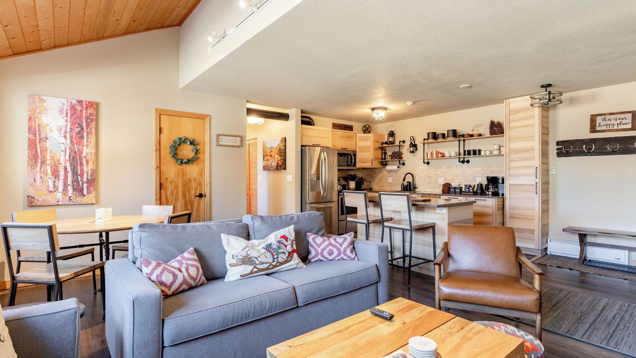 Ein modernes Wohnzimmer mit grauem Sofa, Esstisch aus Holz und einer Küchenzeile mit Holzschränken und einem Kühlschrank.