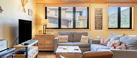 A cozy living room with a sofa, armchair, and coffee table, wooden furniture, a television, and large windows with a view of buildings and mountains.