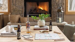 Une pièce chaleureuse avec une cheminée, une table en bois sur laquelle sont posés des livres, des bouteilles et un jeu de société.