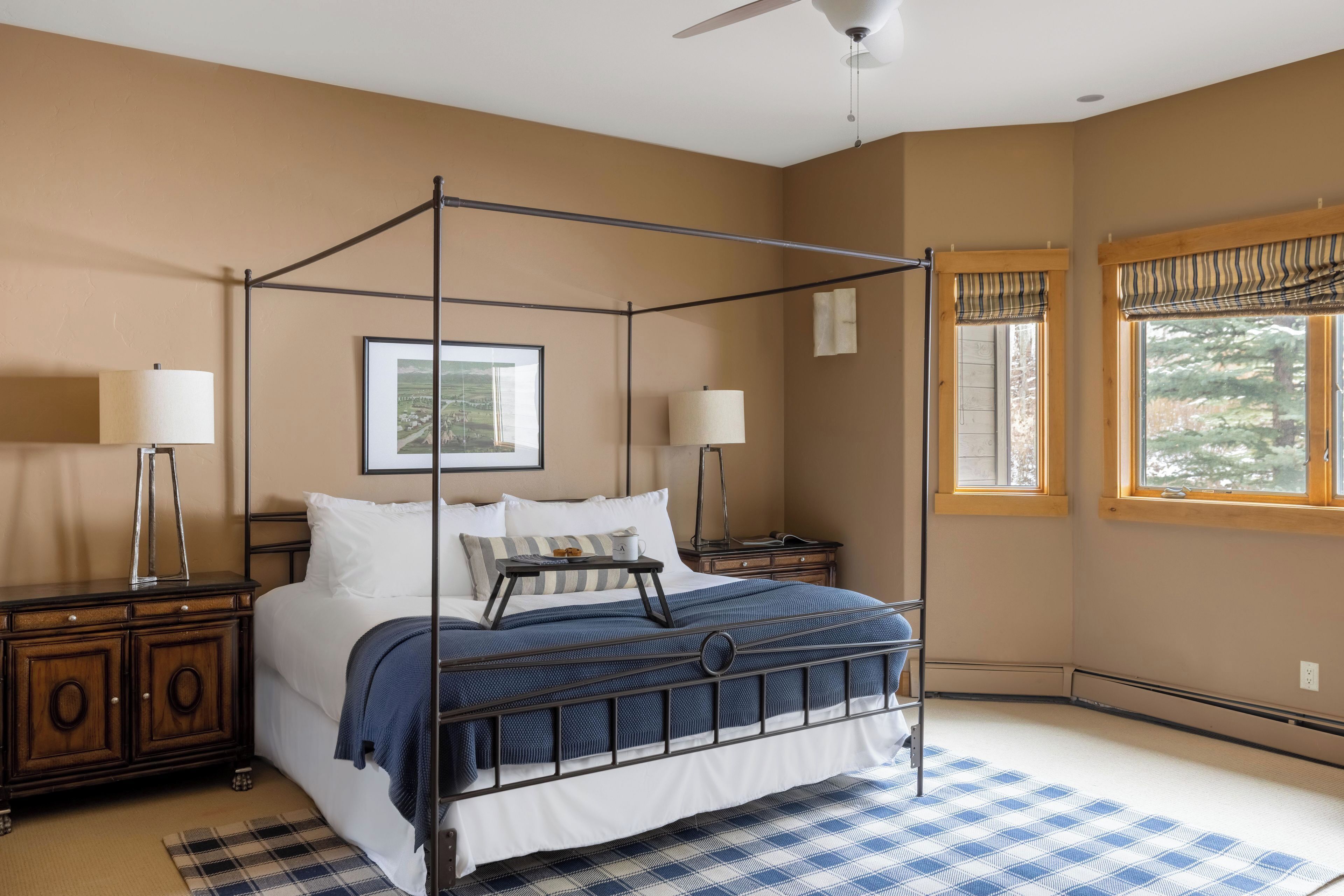 A four-poster bed with a metal frame, two bedside tables, a window with a view of trees, and a ceiling fan.