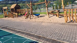 Un terrain de jeux avec un toboggan en bois et des structures d’escalade, le tout situé devant un paysage de maisons et de collines.