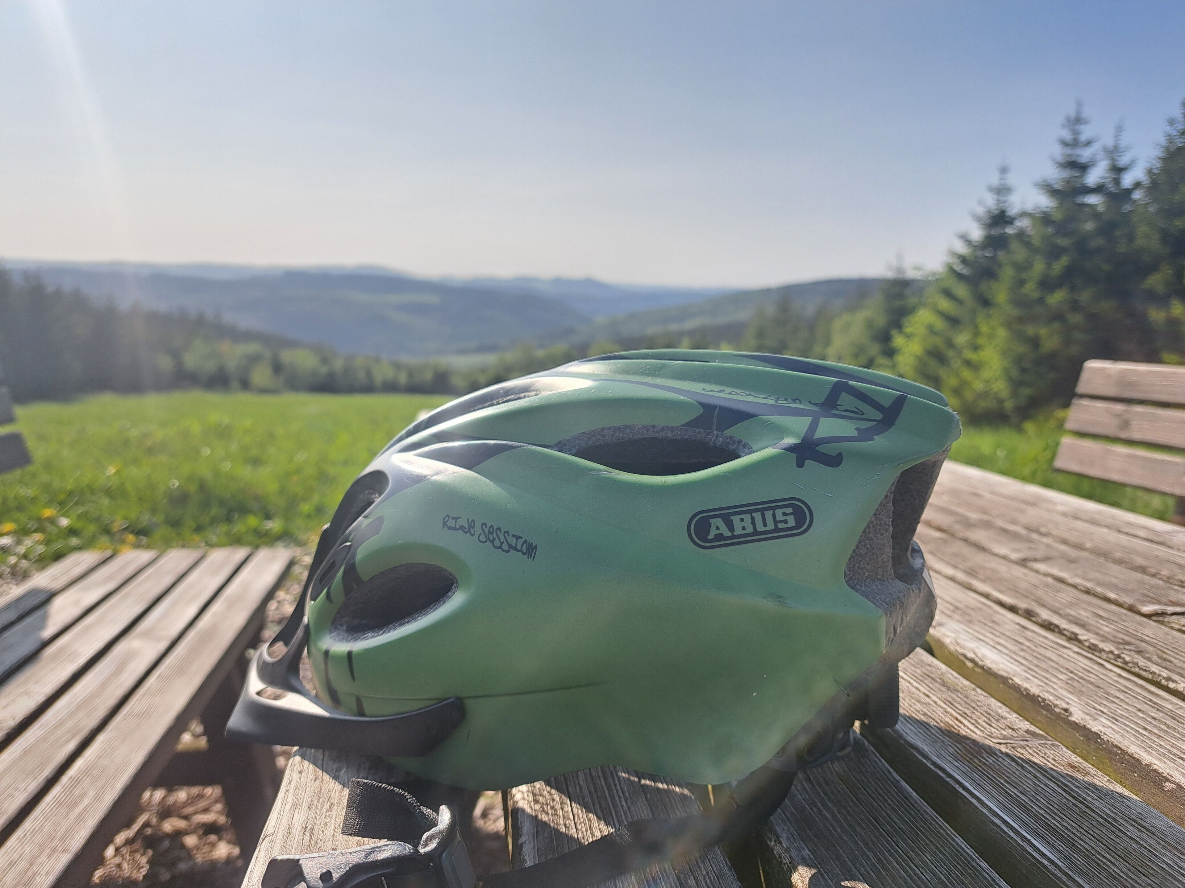 A green bicycle helmet on a wooden bench with a scenic view of a forest and mountains in the background.