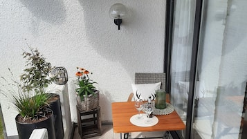 A small outdoor patio with a wooden table, two wicker chairs, potted plants, and a wall-mounted light fixture.