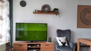 Un salon avec un téléviseur à écran plat affichant le message « PROFITEZ DE LA MUSIQUE », un meuble télé en bois, un fauteuil gris orné d’un coussin à motif de cerf, une horloge murale et une étagère décorative.