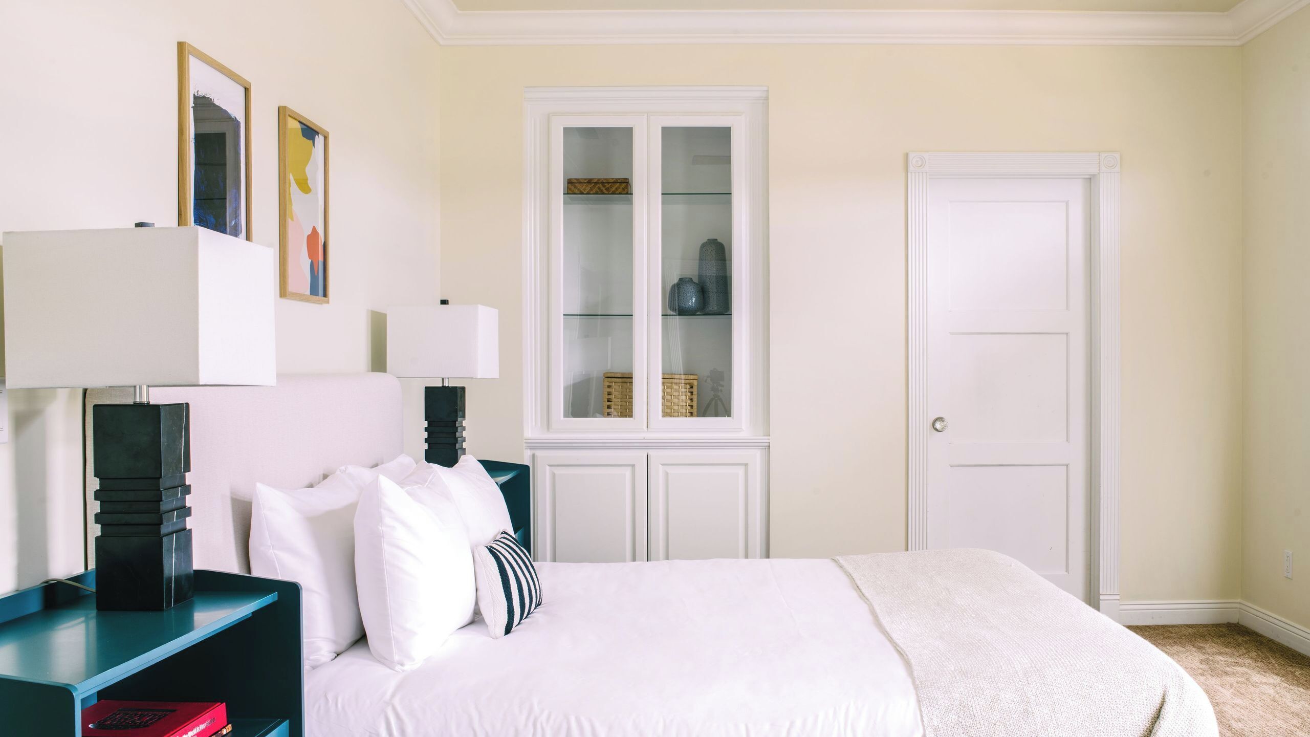 Une chambre à coucher avec un lit, une table de chevet, une lampe, une fenêtre donnant sur une autre pièce et une porte.