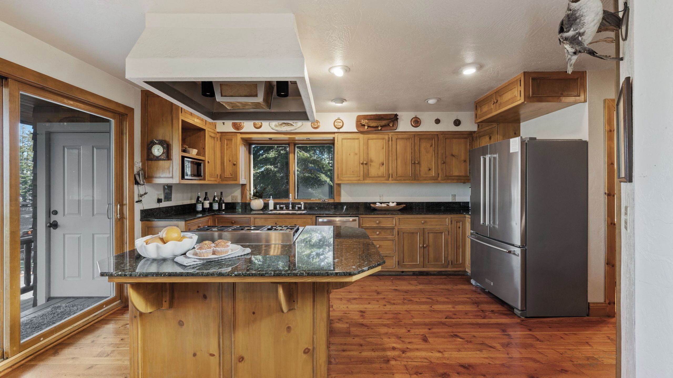 A kitchen with wooden cabinets, a stainless steel refrigerator, and a large island with a bowl of fruit and muffins.