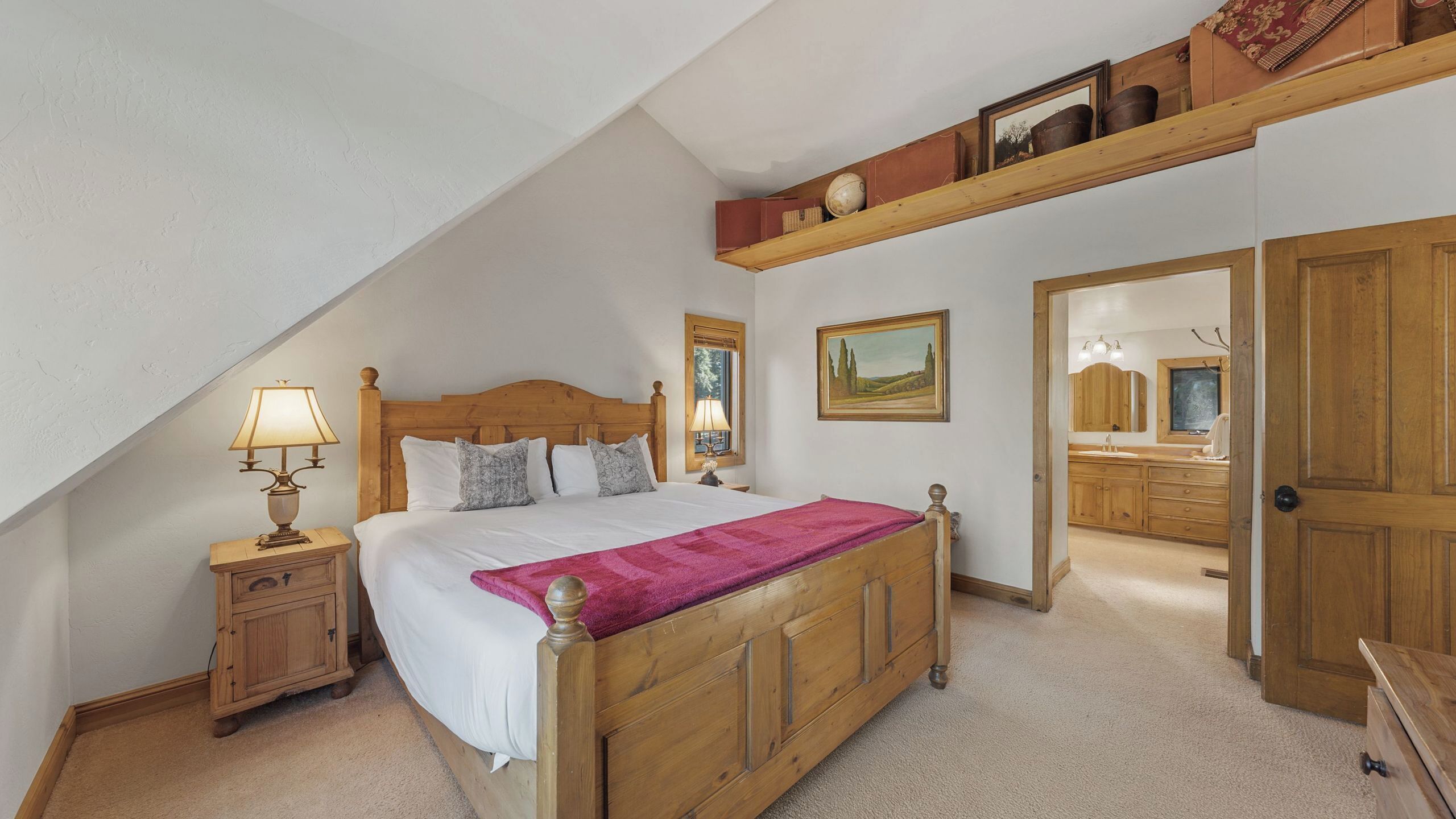 A bedroom with a wooden bed, a nightstand with a lamp, a window, a painting, and a door leading to a bathroom.