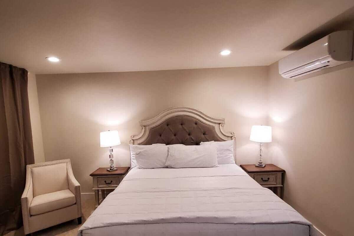 A hotel room with a large bed, two bedside tables with lamps, a chair, an air conditioning unit, and a ceiling light.