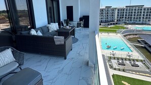 Un espacio exterior moderno con una sala, sillas y una mesa de comedor. Cuenta con una piscina, tumbonas y vistas a un edificio de hotel.