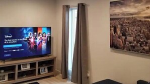 Een woonkamer met een flatscreen-televisie waarop een Disney+-welkomstscherm staat, een plafondventilator, een stadsgezicht schilderij en een houten tv-meubel.