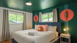 Un lit bien fait, avec une couette blanche et une serviette pliée, une table de chevet avec une lampe, et deux fenêtres avec des rideaux.