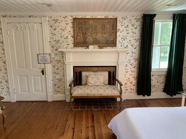 A room with a wooden floor, a white door, a fireplace with a painting above it, and a bed with a white cover.
