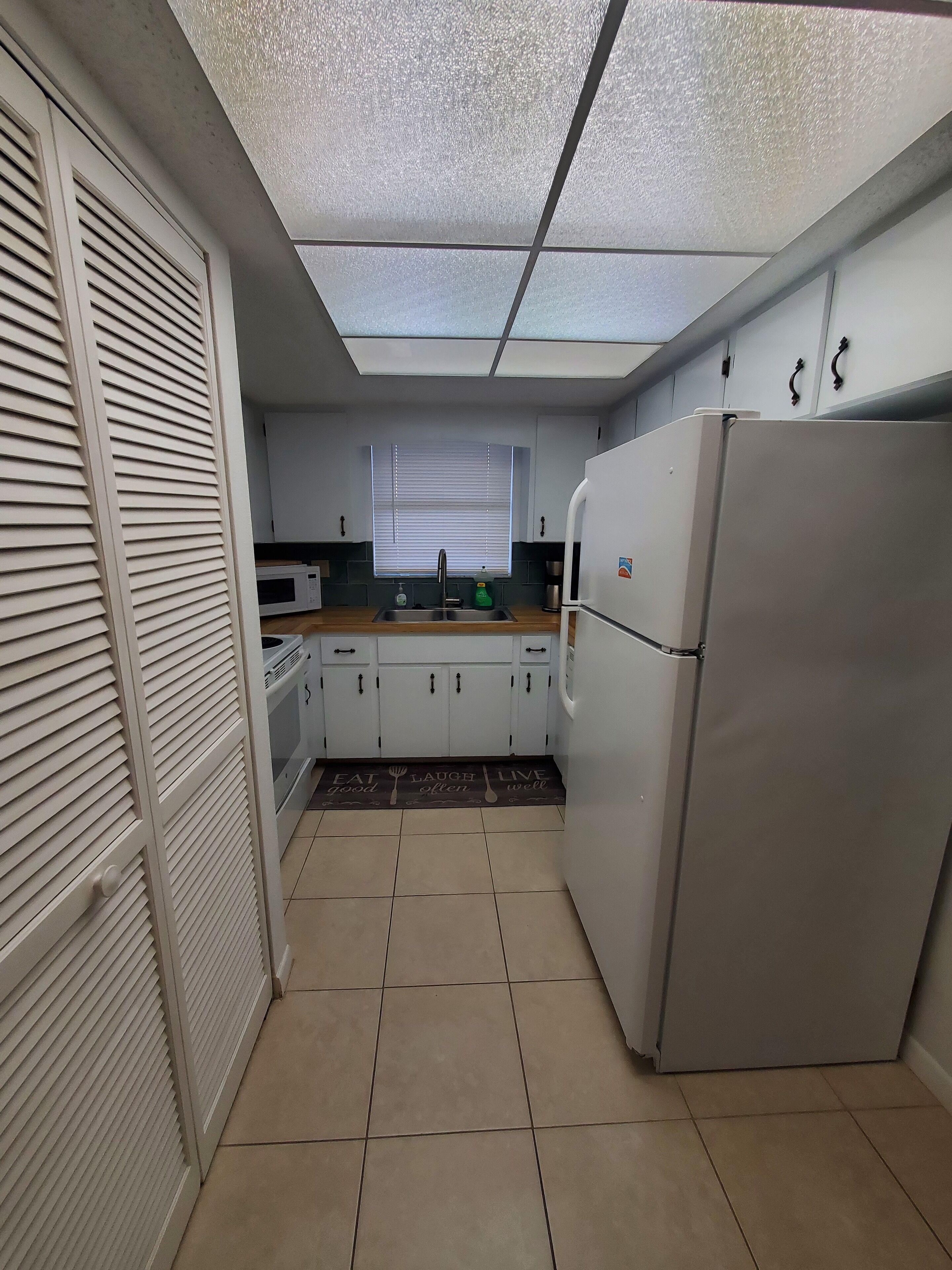Una cocina compacta con armarios blancos, un refrigerador, un microondas y una ventana con persianas.
